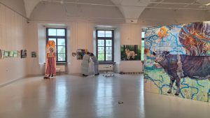 Mākslas galerijas telpā apskatāma laikmetīgās mākslas izstāde ar dažādiem glezniecības darbiem, tostarp lielformāta gleznu ar govs attēlu.