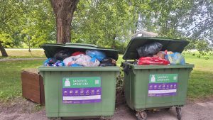 Divi pārpildīti sadzīves atkritumu konteineri zaļā teritorijā.