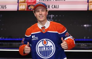 Jaunais hokejists pēc NHL drafta pozē Edmontonas "Oilers" kreklā un cepurē.