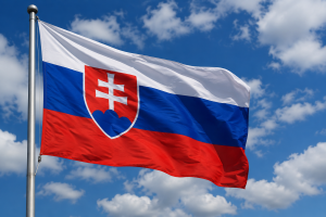 Slovākijas valsts karogs plīvo vējā uz zila debesu fona ar baltiem mākoņiem.