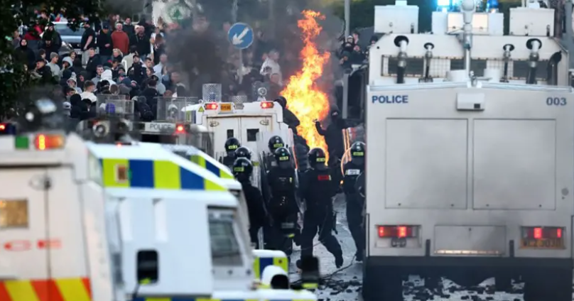 Policijas klātbūtne Belfāstas ielās pretimigrācijas protestu laikā, fonā redzamas liesmas un demonstranti.