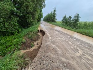 Lietusgāžu izskalots grants ceļš ar bojātu nomali un izveidojušos izskalojumu ceļa malā.