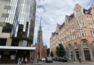 Rīgas centra iela ar vēsturiskām ēkām un baznīcas torni fonā. Ielas malās novietotas automašīnas, bet modernā stikla ēka kontrastē ar apkārtējo vēsturisko apbūvi.