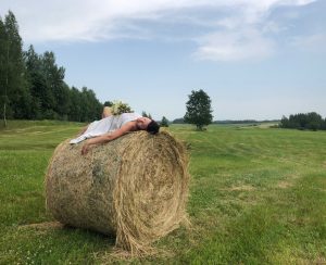 Sieviete baltā kleitā guļ uz siena ruļļa plašā lauku pļavā vasaras saulgriežu noskaņā.