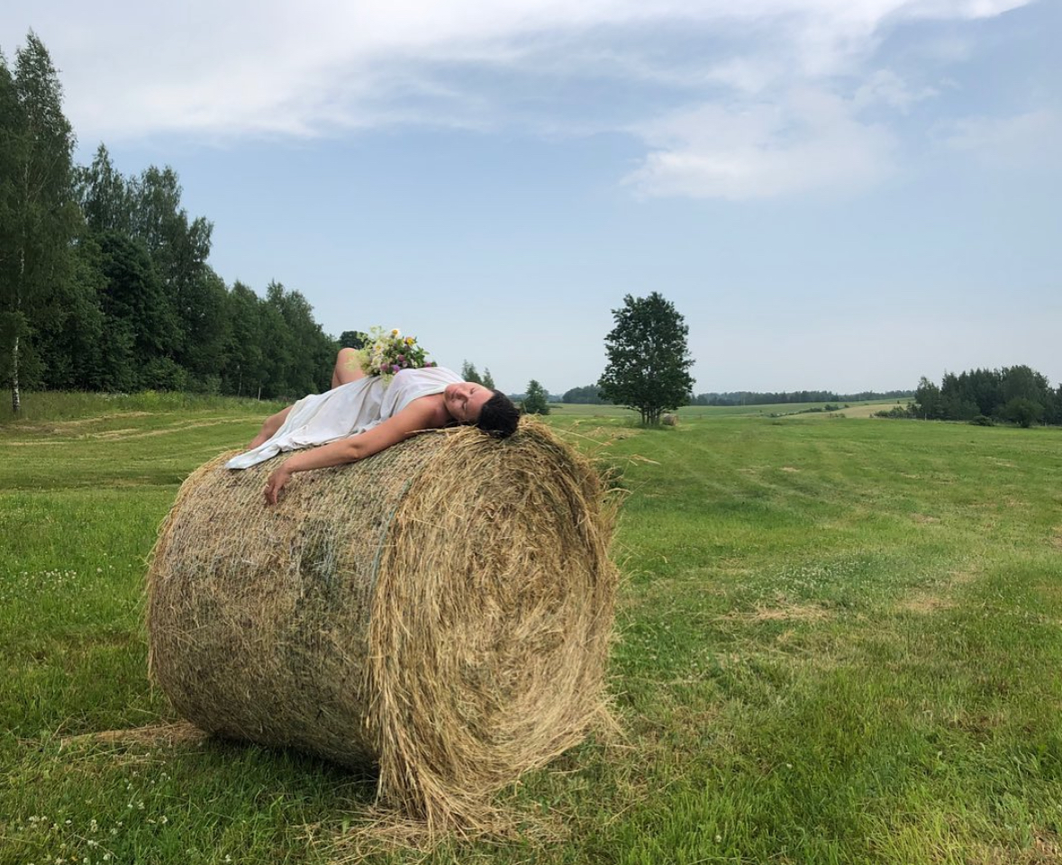 Sieviete baltā kleitā guļ uz siena ruļļa plašā lauku pļavā vasaras saulgriežu noskaņā.