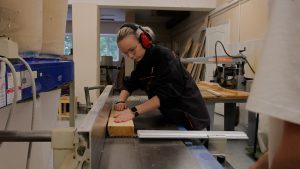 Audzēkne profesionālās izglītības darbnīcā apgūst kokapstrādes prasmes, strādājot ar ēveli un ievērojot darba drošības prasības.
