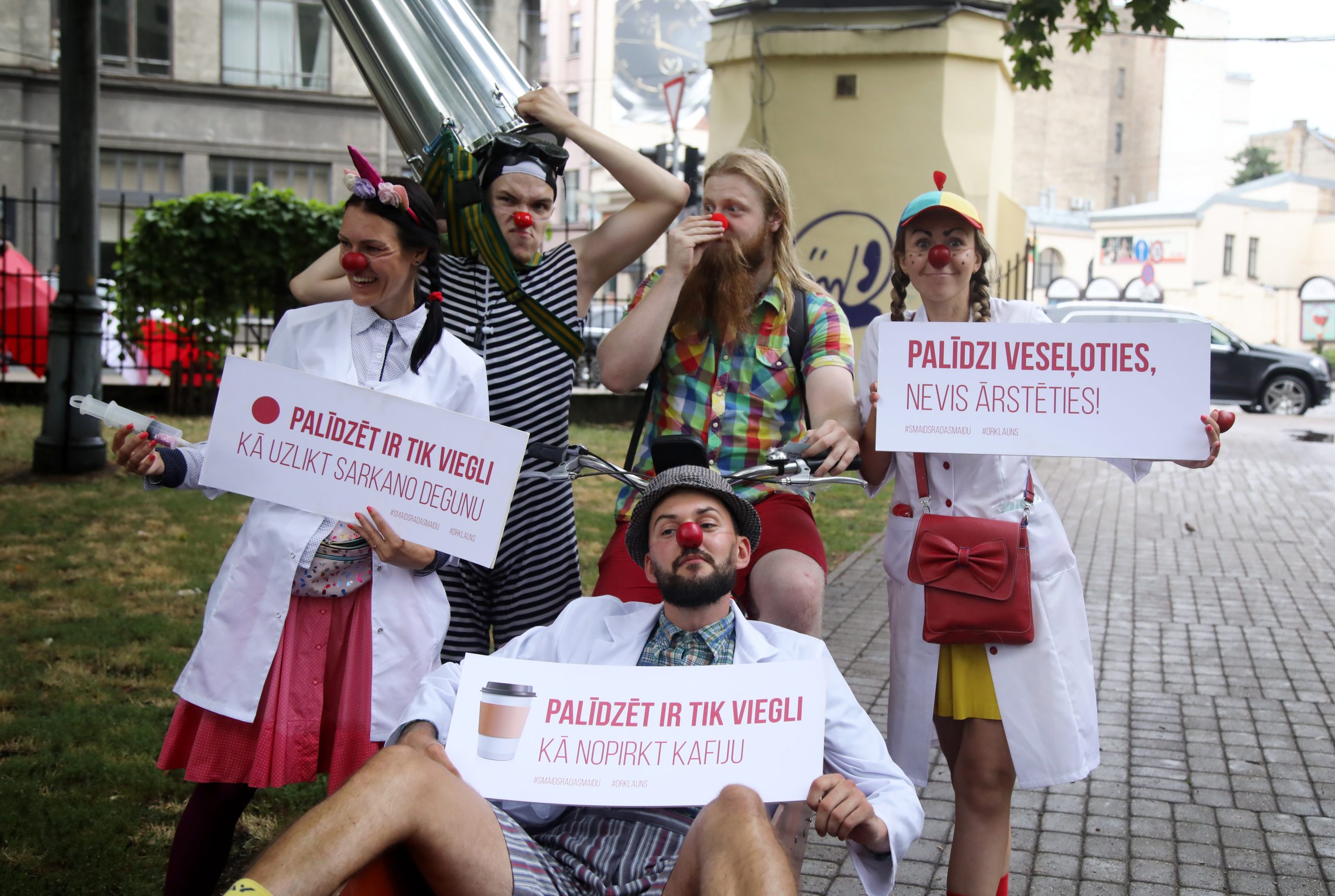 Dr.Klauns kustības pārstāvji ar sarkanajiem klaunu deguniem Vērmanes dārzā tur plakātus vasaras kampaņas atklāšanas pasākumā.