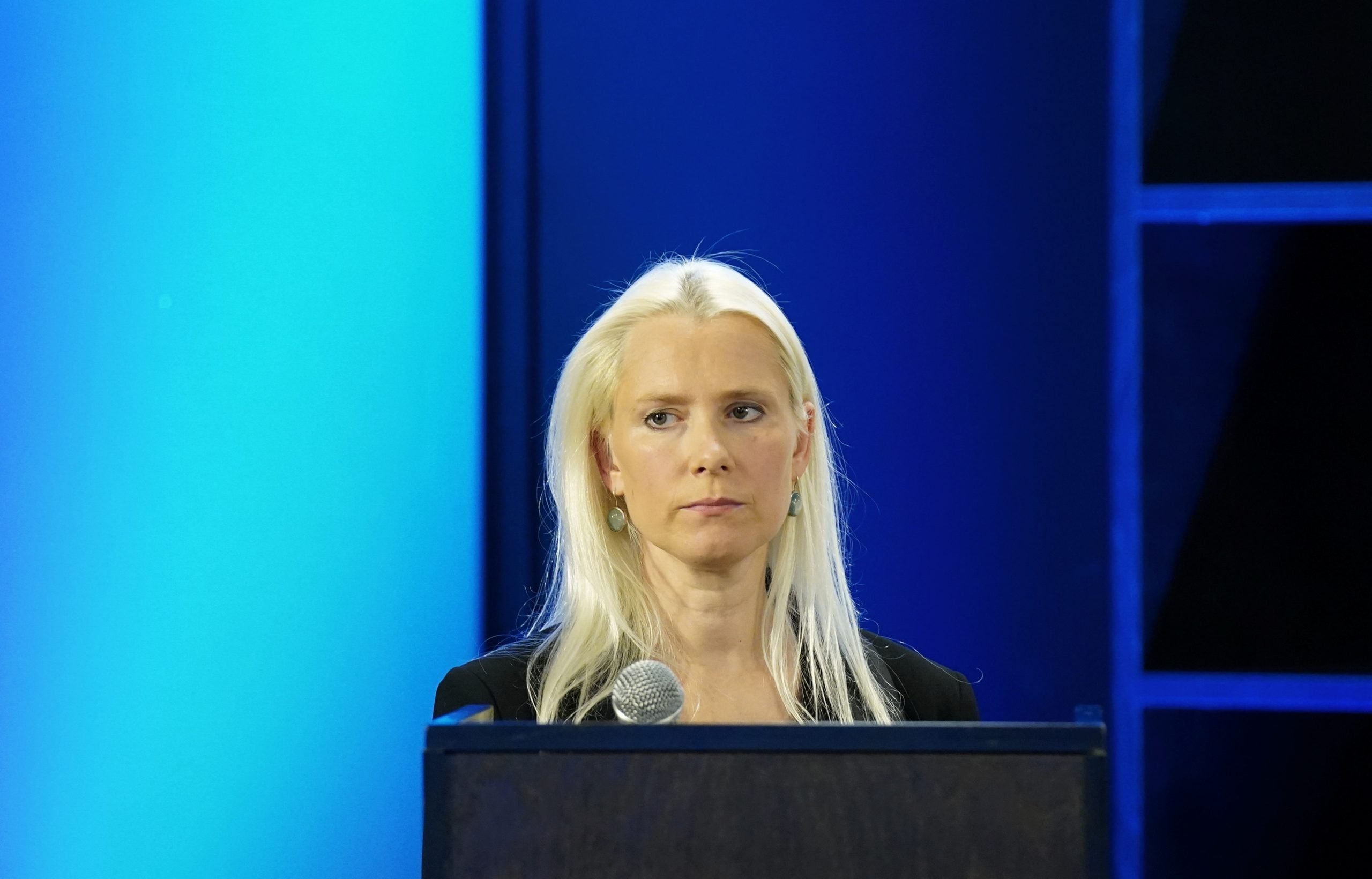 Aurēlija Ieva Druviete uzstājas pie tribīnes publiskā pasākumā vai konferencē, fonā redzams zils apgaismojums.