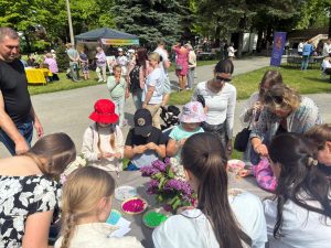 Bērni un pieaugušie piedalās radošajā darbnīcā parkā, veidojot rotaslietas no krāsainām pērlītēm.