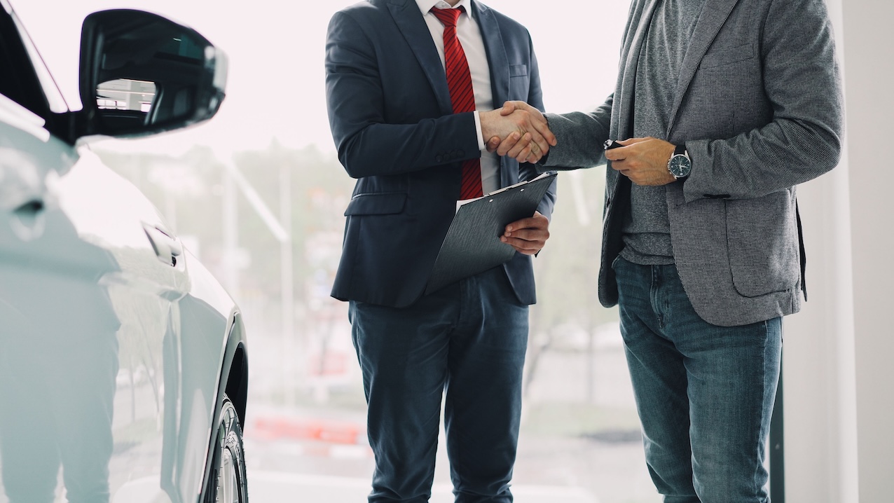 Autosalona darbinieks un klients paspiež rokas pie automašīnas pēc līguma noslēgšanas.