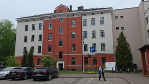 Daudzstāvu Jelgavas Klīnikas ēka ar autostāvvietu priekšplānā.