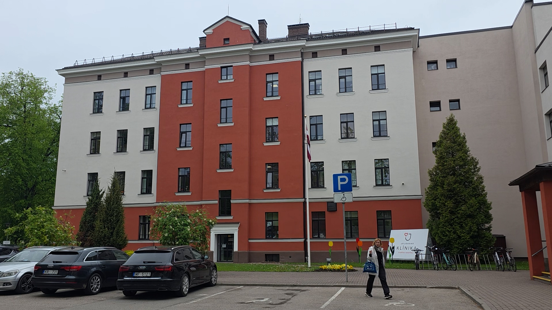 Daudzstāvu Jelgavas Klīnikas ēka ar autostāvvietu priekšplānā.