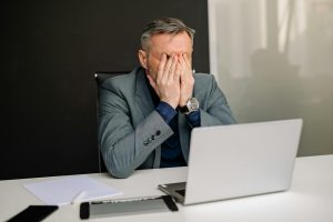 Vīrietis biroja vidē sēž pie klēpjdatora un aizsedz seju ar rokām, paužot stresu, nogurumu vai emocionālu pārslodzi