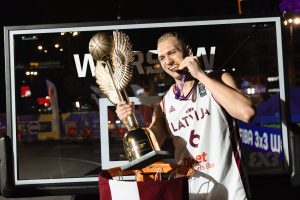 Latvijas 3x3 basketbola izlases spēlētājs pozē ar Pasaules kausa trofeju un zelta medaļu pēc uzvaras turnīrā.