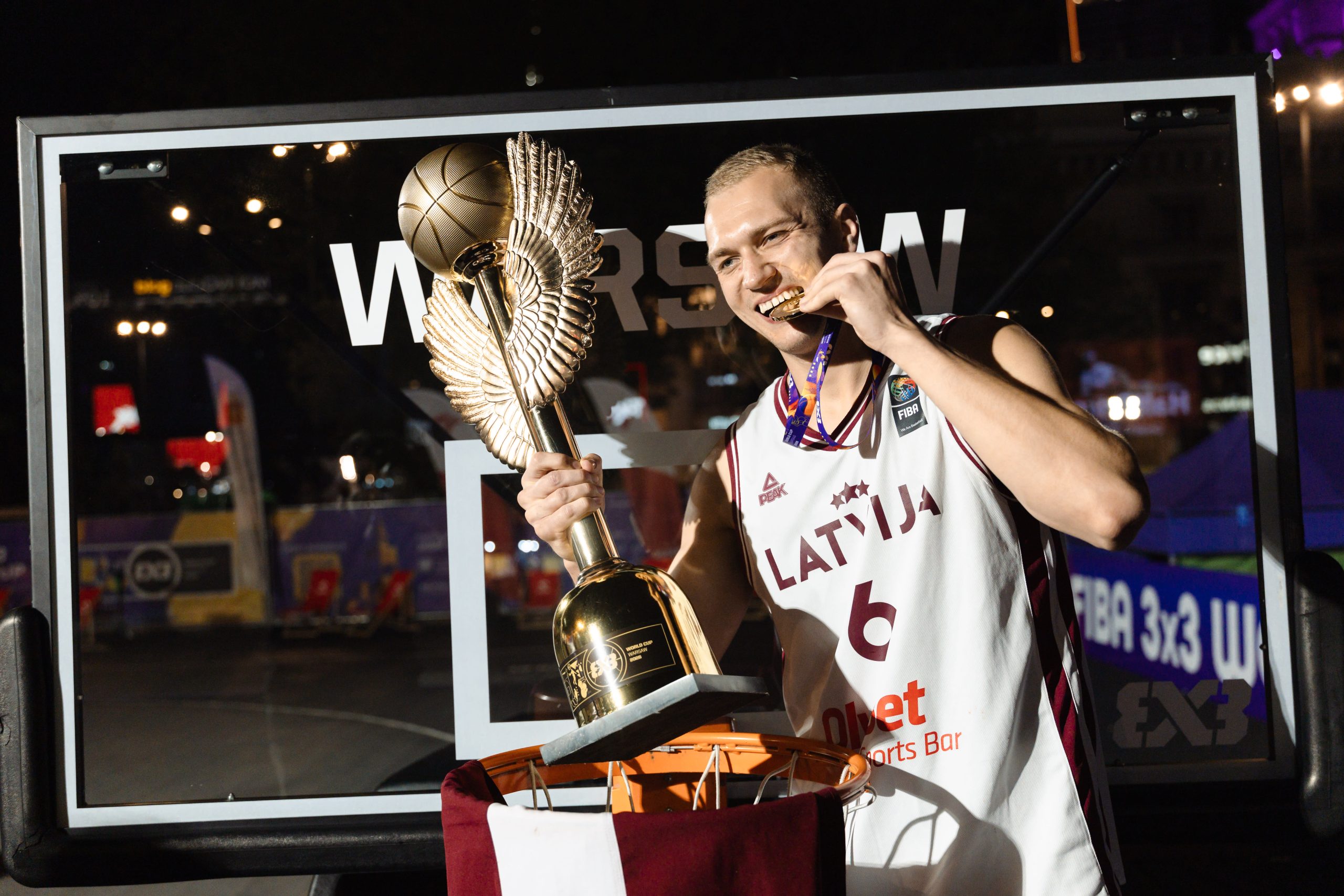Latvijas 3x3 basketbola izlases spēlētājs pozē ar Pasaules kausa trofeju un zelta medaļu pēc uzvaras turnīrā.