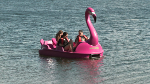 Cilvēki brauc ar rozā flamingo formas ūdens velosipēdu.