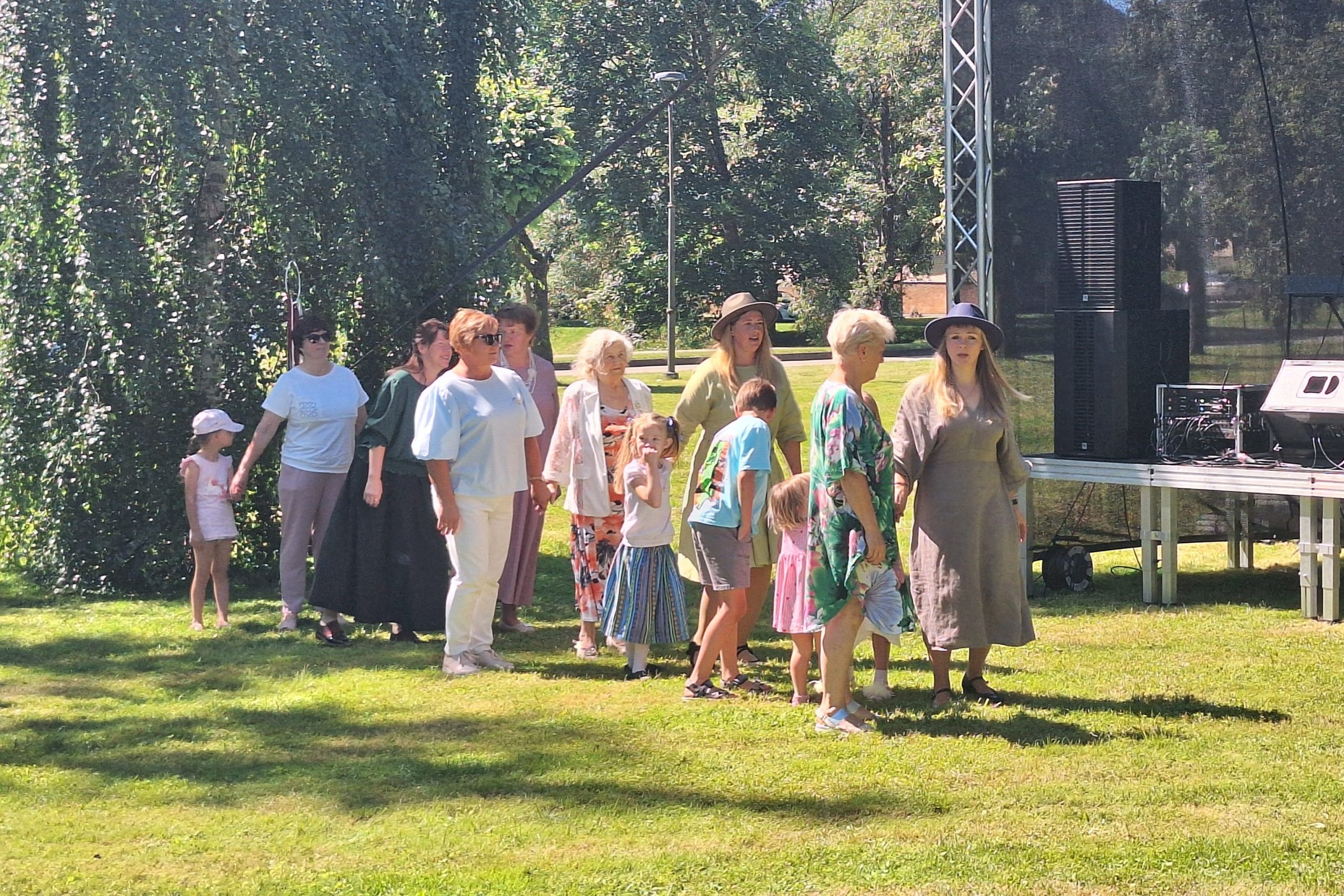 Cilvēku grupa, tostarp bērni, pulcējas pie brīvdabas skatuves parkā, gatavojoties pasākumam.