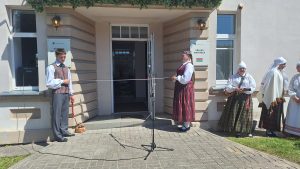 Pie ēkas ieejas tradicionālajos tautastērpos tērpti cilvēki gatavojas lentes pārgriešanai.