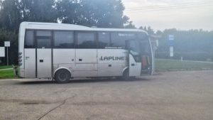 Balts autobuss stāv autobusu pieturā ar atvērtām priekšējām durvīm.