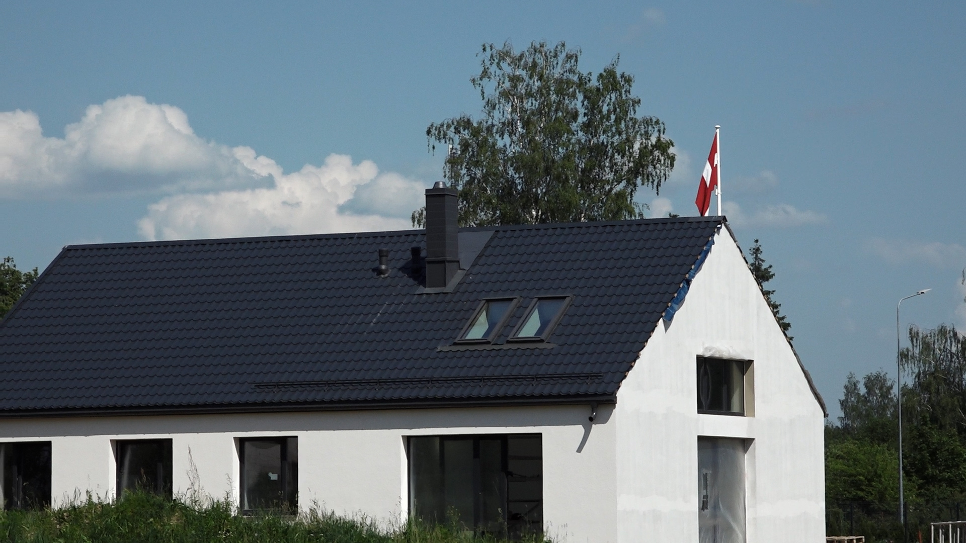 Jaunuzcelta privātmāja ar tumšu jumtu un Latvijas karogu pie ēkas.