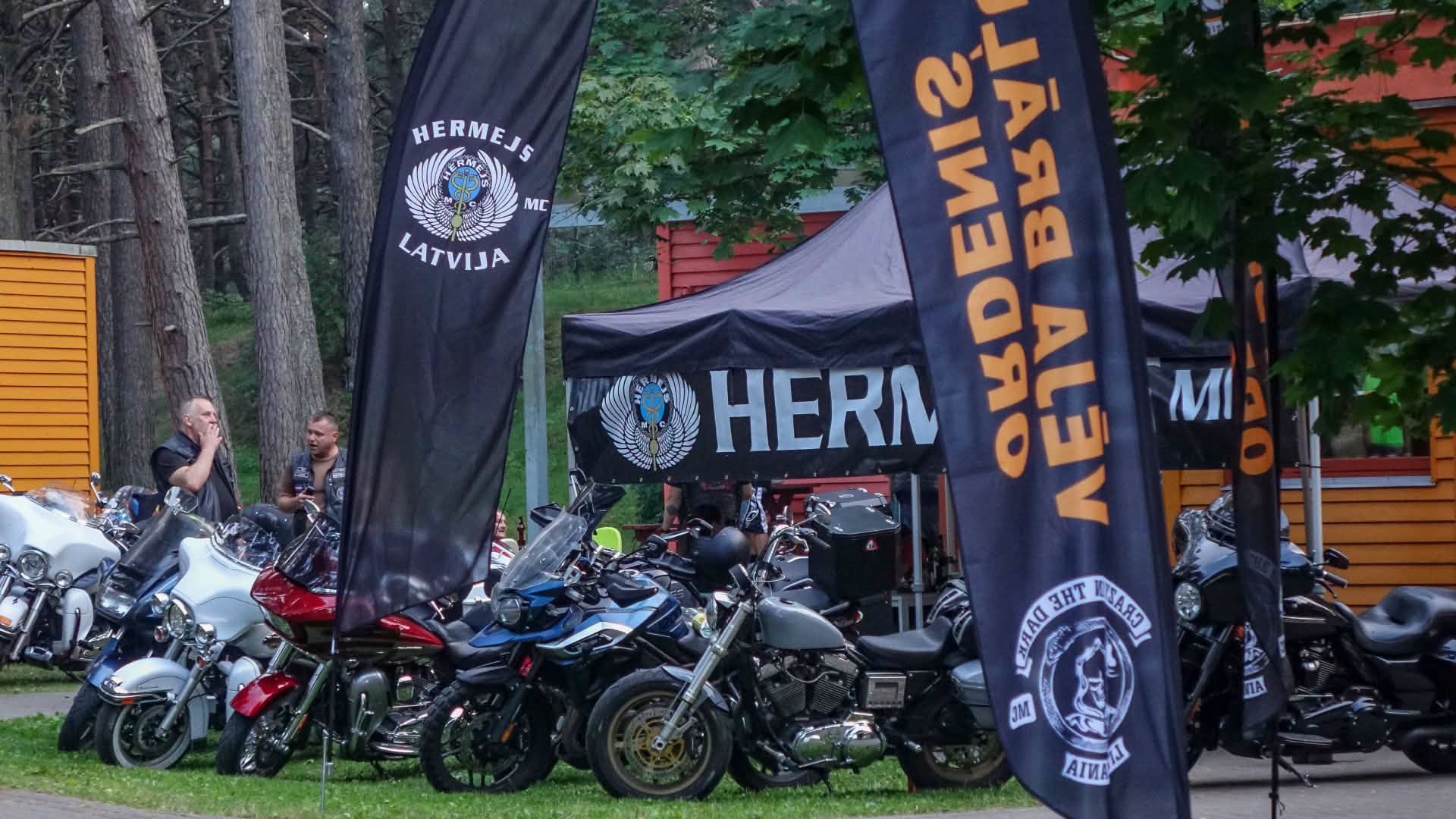 Motociklu saieta teritorijā pie teltīm novietoti vairāki motocikli un plīvo motociklistu klubu karogi.