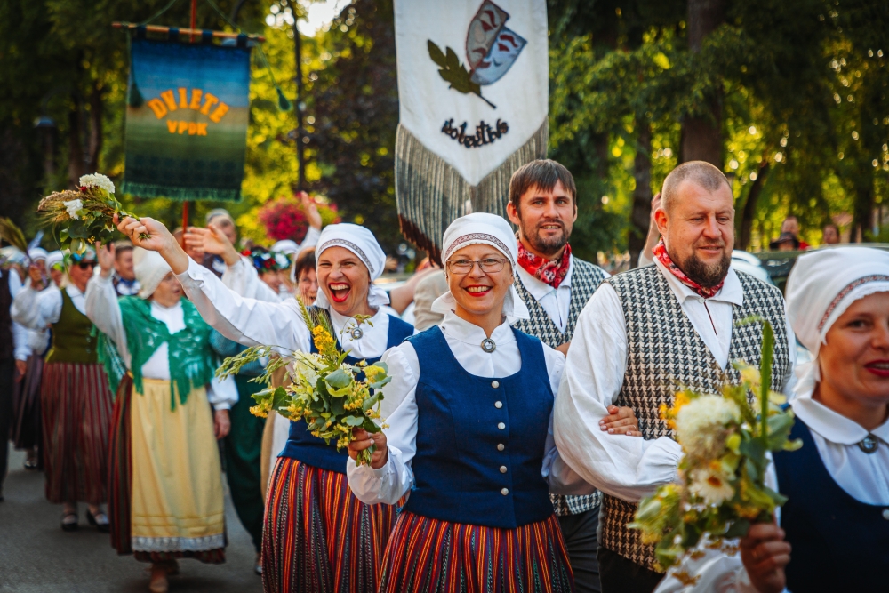 Cilvēki tautastērpos dodas svētku gājienā, rokās turot ziedus un ozollapu pušķus.