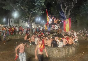 Cilvēku pūlis pulcējies ap strūklaku Spānijas pilsētā, daļa svinētāju uzkāpuši uz strūklakas malas un augšējās konstrukcijas, vicinot Spānijas karogus.