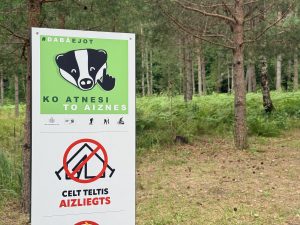 Informatīva zīme mežā aicina apmeklētājus aiznest savus atkritumus un informē, ka telšu celšana ir aizliegta.