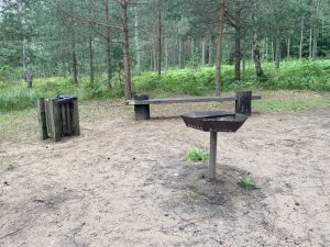 Atpūtas vieta mežā ar grilu, soliņu un atkritumu urnu dabas parka teritorijā.