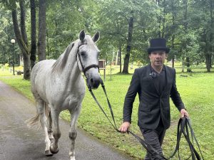 Zirgu treneris Sandis Ģērmanis ved baltu zirgu pa parka celiņu.