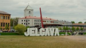 Jelgavas pilsētvide ar lielajiem “Jelgava” burtiem, ēkām un gājēju tiltu.