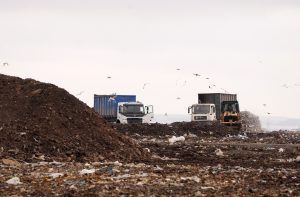 Atkritumu poligonā Getliņi redzamas kravas automašīnas un buldozers starp atkritumu kaudzēm.