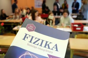 Fizikas skolotāja rokasgrāmata klasē ar skolēniem fonā.