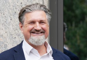 Smaidīgs vīrietis tumšā žaketē un baltā kreklā pie ēkas fasādes.