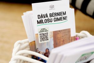 Informatīvi bukleti par iespēju kļūt par uzņemošo ģimeni ievietoti baltā pītā grozā.