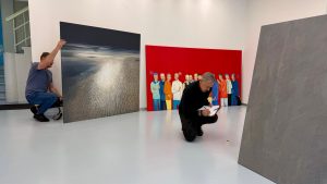 Mākslas galerijā "Manss" tiek iekārtota vasaras izstāde – darbinieki izvieto lielformāta gleznas ekspozīcijas zālē.