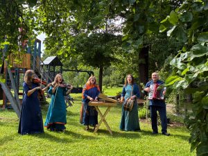 Igauņu ģimene muzicē dārzā – ģimenes locekļi spēlē vijoles, kokli, bungas un akordeonu.