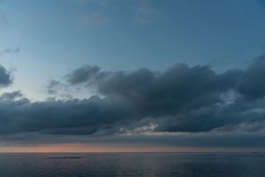 Tumši negaisa mākoņi virs mierīgas jūras saulrietā, pie horizonta redzama kuģa siluets.