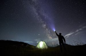 Cilvēks naktī pie telts ar lukturīti izgaismo zvaigžņotās debesis un Piena Ceļu.
