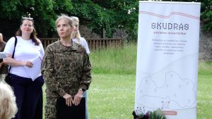Biedrības "Sievietes aizsardzībai Skudras" pasākumā Cēsīs uzstājas instruktore militārā formas tērpā, fonā redzams biedrības informatīvais stends.