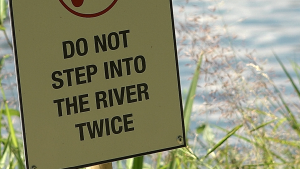 Brīdinājuma zīme ar uzrakstu "Do not step into the river twice" pie ūdenstilpes Strenčos.