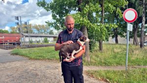 Vīrietis tur rokās mazu suni pie dzelzceļa pārbrauktuves