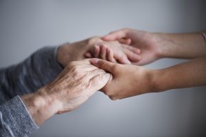 Jaunāks cilvēks maigi tur seniora rokas, simbolizējot aprūpi, atbalstu un līdzcilvēku rūpes.