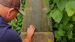 Vīrietis aplūko sena krucifiksa pamatnē iegravētu uzrakstu Dagdas pusē