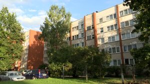 Daudzdzīvokļu dzīvojamā māja ar nolietotu fasādi, ko ieskauj koki un apstādījumi.