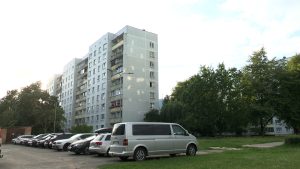 Daudzdzīvokļu dzīvojamā māja ar autostāvvietu un zaļo zonu priekšplānā.