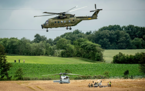 Militārais helikopters lido virs lauka, kur redzams vēl viens helikopters un cilvēki