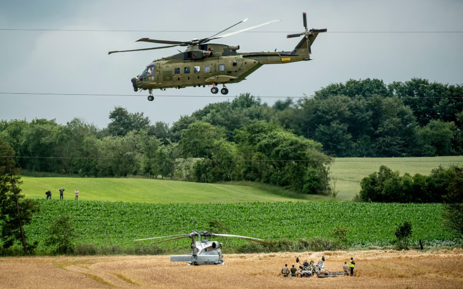 Militārais helikopters lido virs lauka, kur redzams vēl viens helikopters un cilvēki