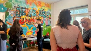 Jaunieši un pieaugušie pulcējas pie galda ar uzkodām Jēkabpils Jauniešu mājas iekštelpās, kur pie sienas ir liels, krāsains grafiti zīmējums.