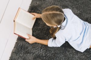 Meitene lasa grāmatu, guļot uz paklāja